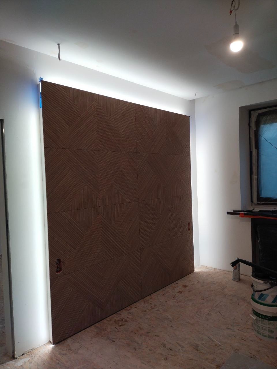 Panel ścienny z geometrycznym wzorem w odcieniach brązu, podświetlony listwą LED, tworzący nowoczesny akcent w jasnym wnętrzu. Widoczne detale wykończenia.