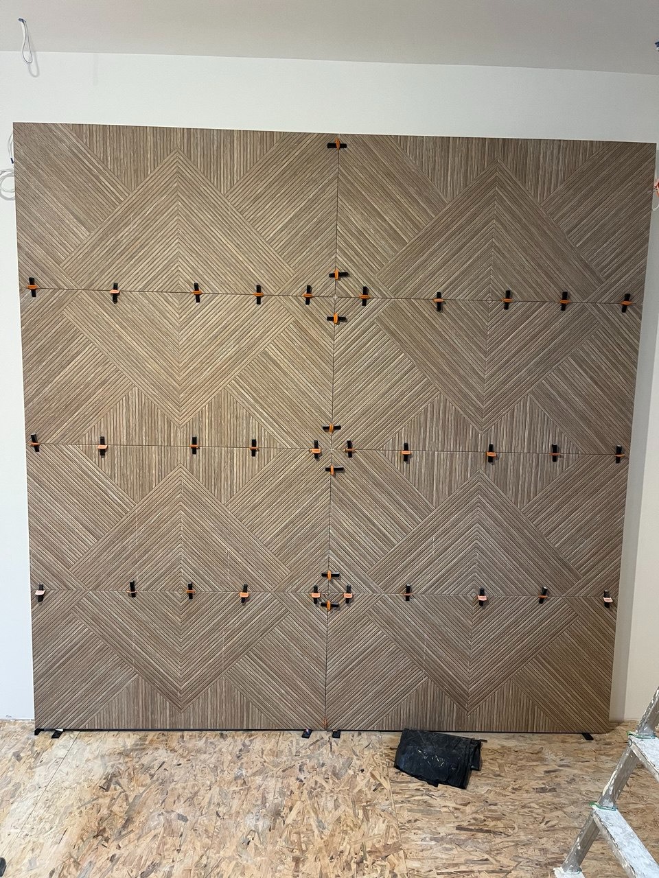 Układanie dekoracyjnych, brązowych płytek ściennych z użyciem klipsów poziomujących. Widoczna drabina i podłoga w trakcie prac wykończeniowych.