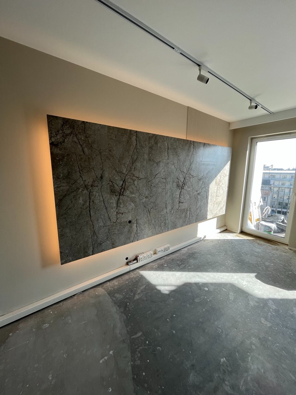 Wykończony fragment salonu z podświetlaną ścianą z płytek imitujących marmur, listwami dekoracyjnymi i oświetleniem szynowym. Widoczne gniazdka elektryczne.