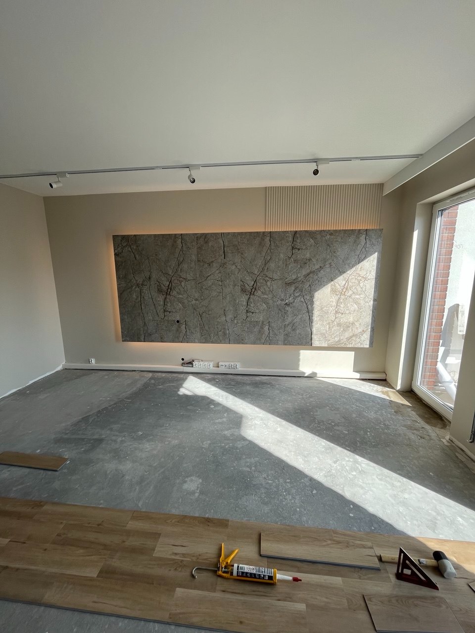 Wnętrze w trakcie wykańczania: panele podłogowe, klej, poziomica. Na ścianie dekoracyjny panel z podświetleniem LED. Widoczne listwy przypodłogowe i oświetlenie sufitowe.