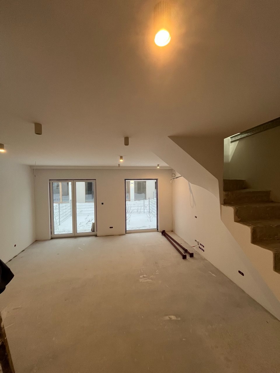 Wnętrze w stanie deweloperskim z widocznymi schodami, oknami balkonowymi i punktami oświetleniowymi na suficie. Podłoga surowa, ściany przygotowane do malowania.