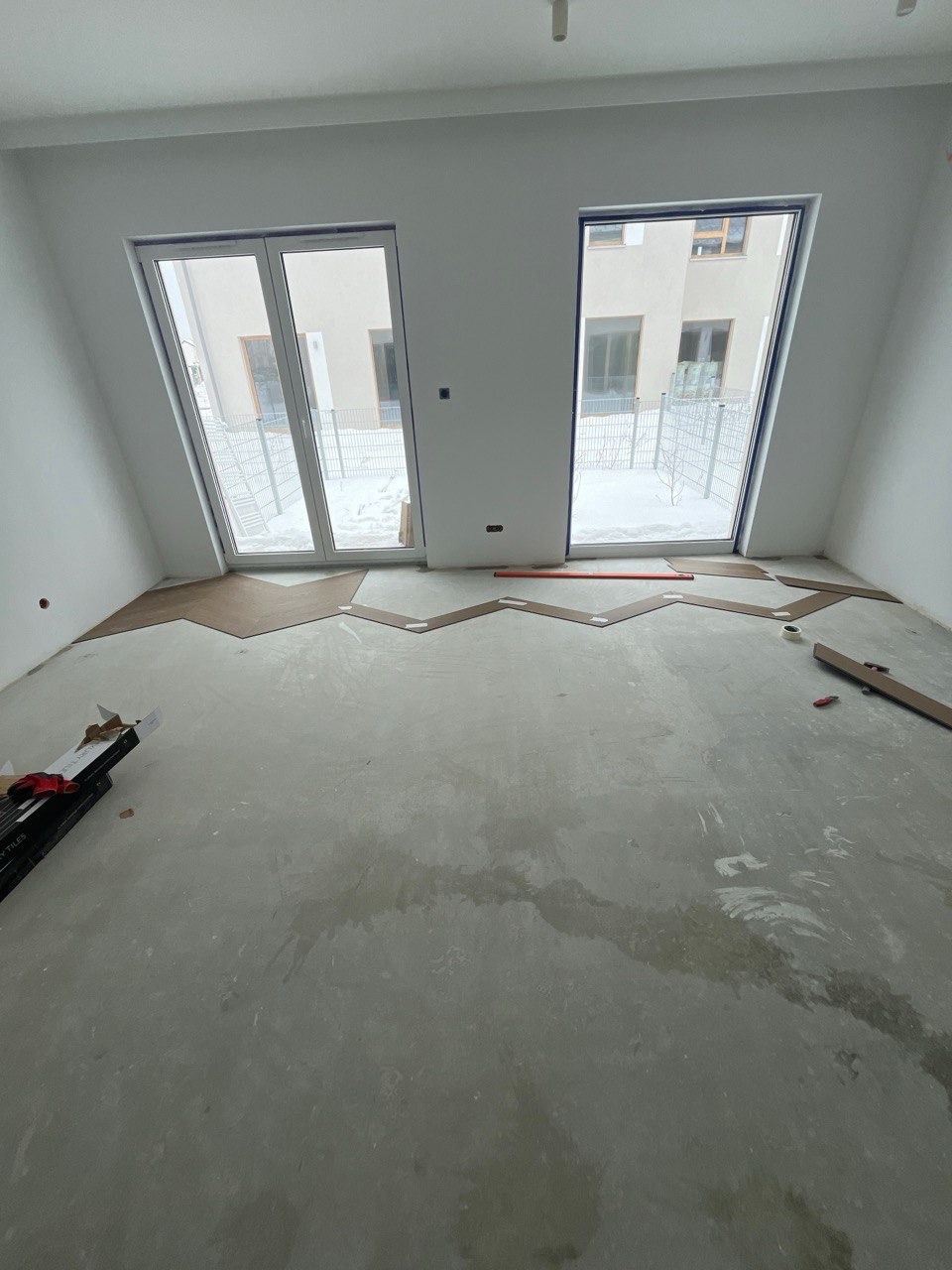 Przygotowanie do montażu podłogi w jasnym pokoju z dwoma oknami. Widoczne płyty podłogowe ułożone częściowo na surowym podłożu. Etap prac wykończeniowych.