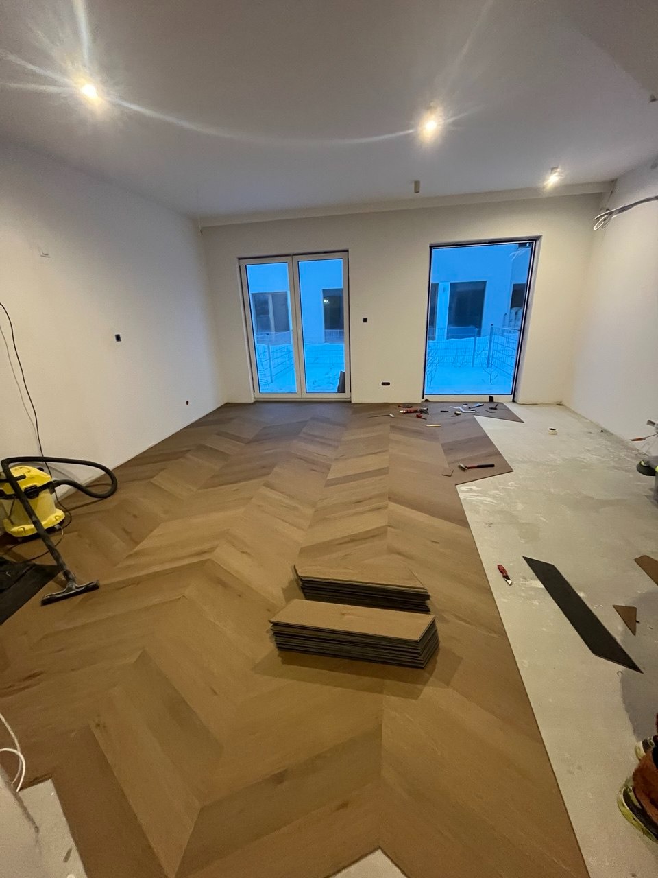 Układanie parkietu w jodełkę w jasnym wnętrzu. Częściowo ułożona podłoga, widoczne paczki paneli i odkurzacz. Dwa okna balkonowe w tle.