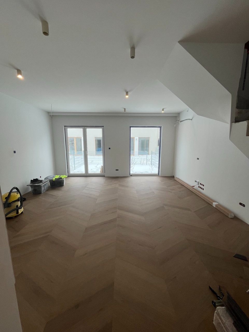 Ukończona podłoga z parkietu w jodełkę w jasnym wnętrzu z oknami balkonowymi w Warszawie. Widoczne narzędzia i akcesoria. Nowoczesne oświetlenie sufitowe.