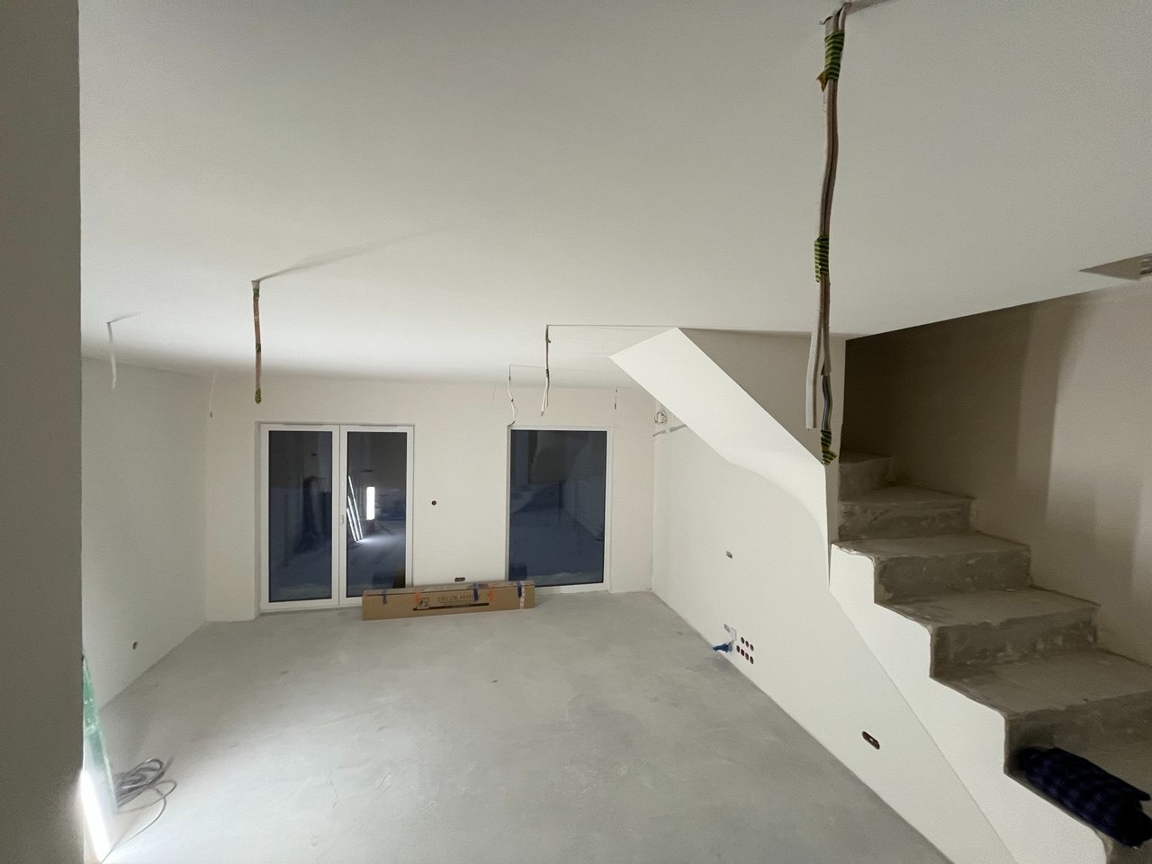 Wnętrze w trakcie wykończenia: surowe schody, gładkie ściany, okna balkonowe, wystające przewody elektryczne. Podłoga wylewka, karton na podłodze.