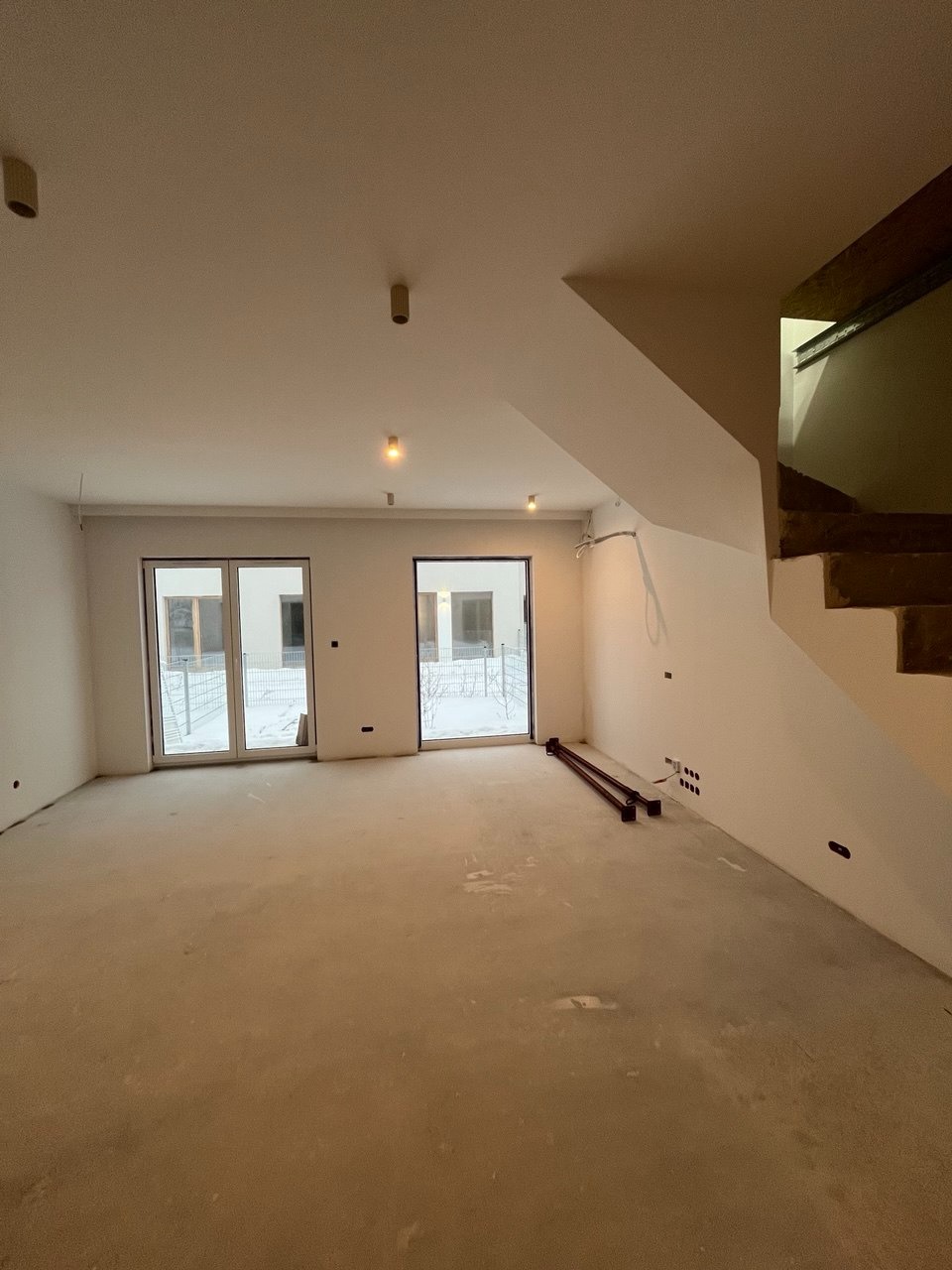 Surowe wnętrze z dwoma oknami balkonowymi i schodami w trakcie wykończenia. Widoczne przygotowane podłoże i instalacje elektryczne. Minimalistyczny styl.