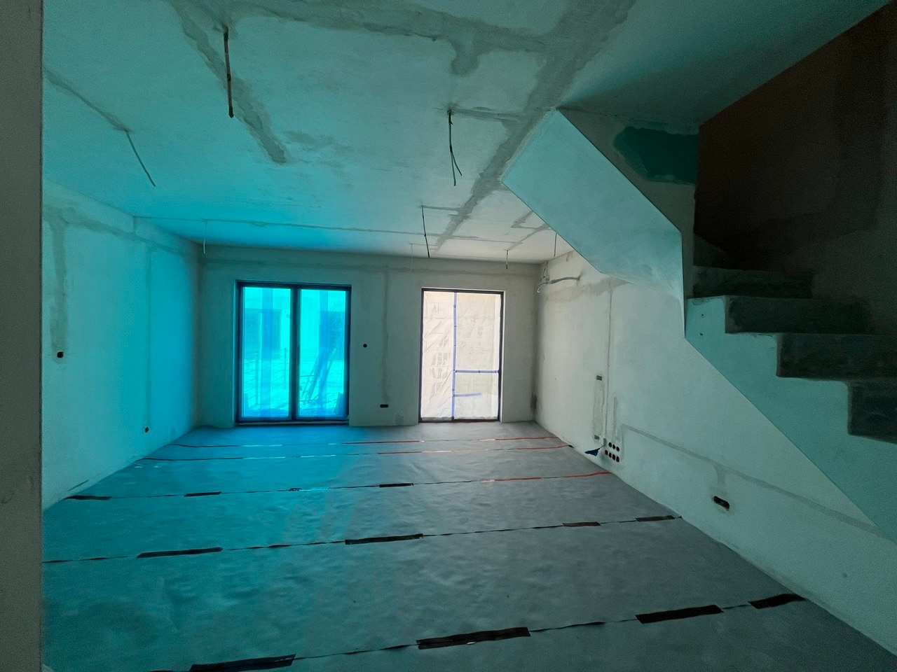 Wnętrze w stanie deweloperskim z zabezpieczoną podłogą, widoczne okna z folią ochronną i surowe schody. Ściany przygotowane do dalszych prac wykończeniowych.