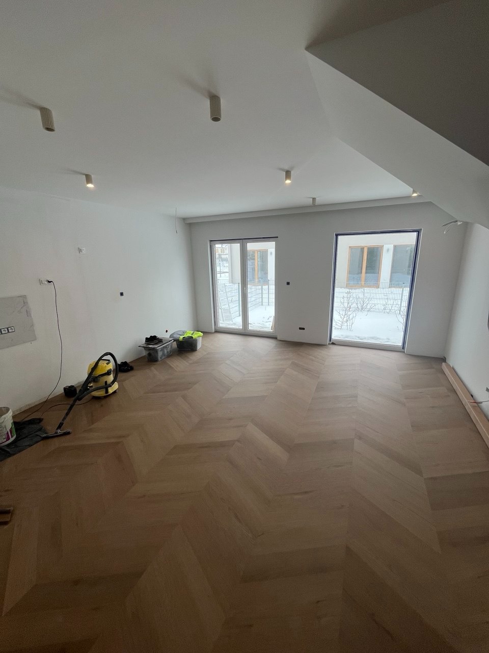 Jasne, parkietowe wykończenie podłogi w jodełkę francuską w przestronnym, białym wnętrzu z dużymi oknami. Widoczny odkurzacz i akcesoria malarskie.