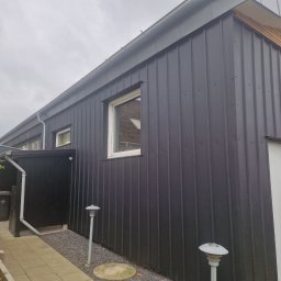 Łukasz Słonina - Nowoczesny budynek z ciemną, pionową blachą trapezową na elewacji, szara rynna, biała stolarka okienna i drzwiowa, oświetlenie zewnętrzne w formie lamp na słupkach.