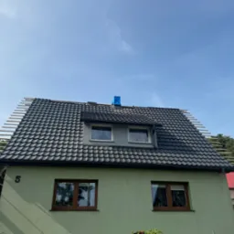 Dom z nowym, czarnym dachem z dachówki ceramicznej w trakcie montażu, widoczne łaty dachowe i zabezpieczony komin niebieską folią.