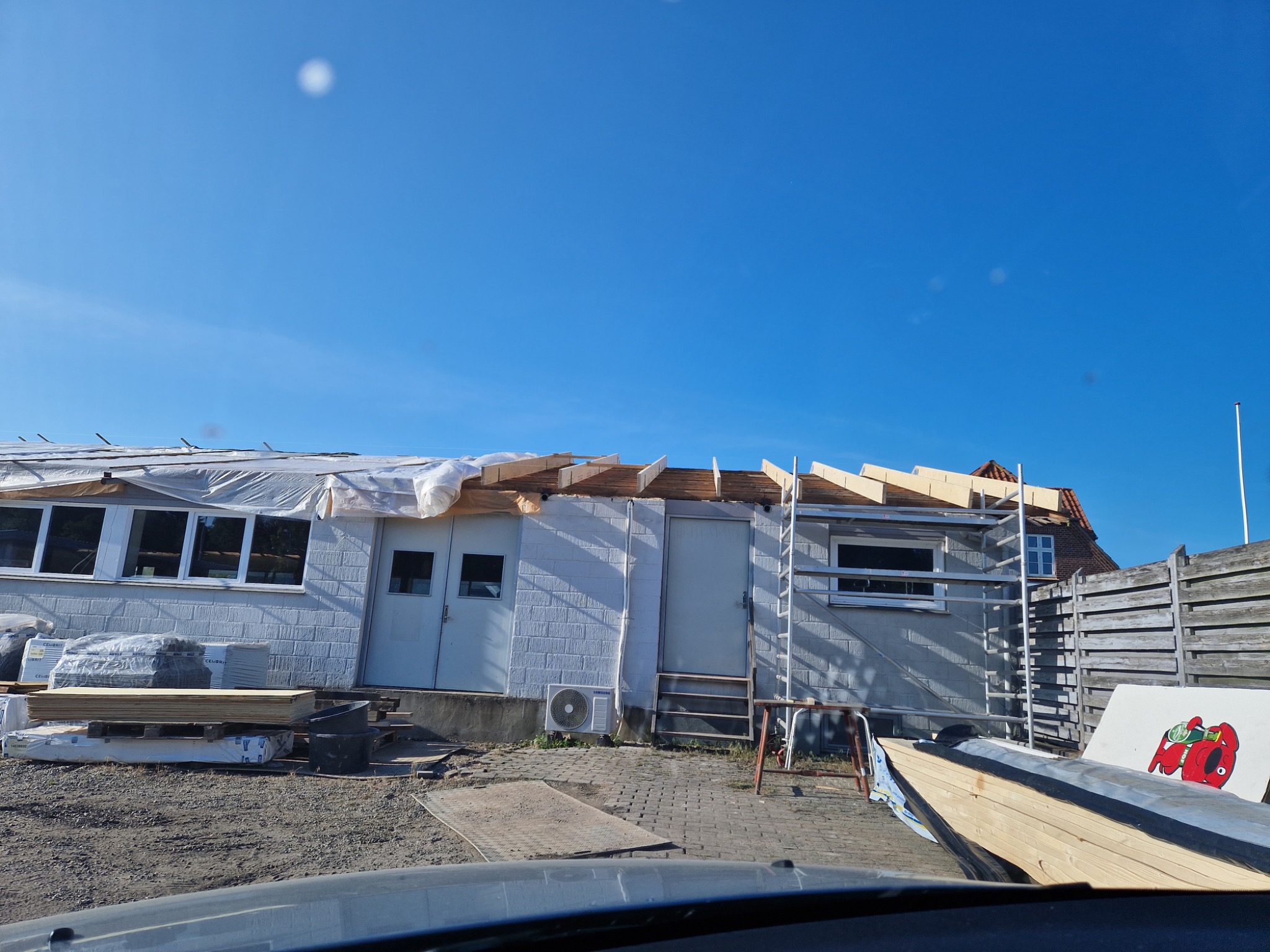 Budynek w trakcie remontu dachu, widoczne drewniane elementy konstrukcyjne, częściowo przykryte folią, z boku budynku leżą materiały budowlane.