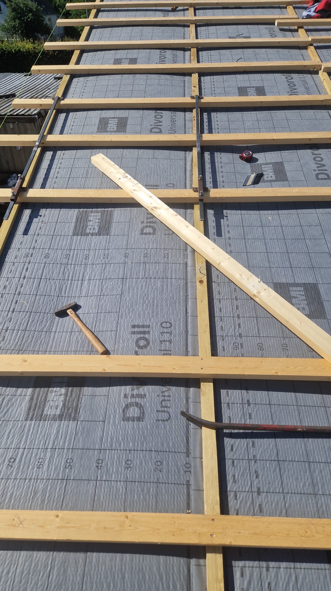 Konstrukcja drewniana dachu pokryta membraną z nadrukowaną siatką i numeracją, widoczne narzędzia: młotek, miarka i łom.