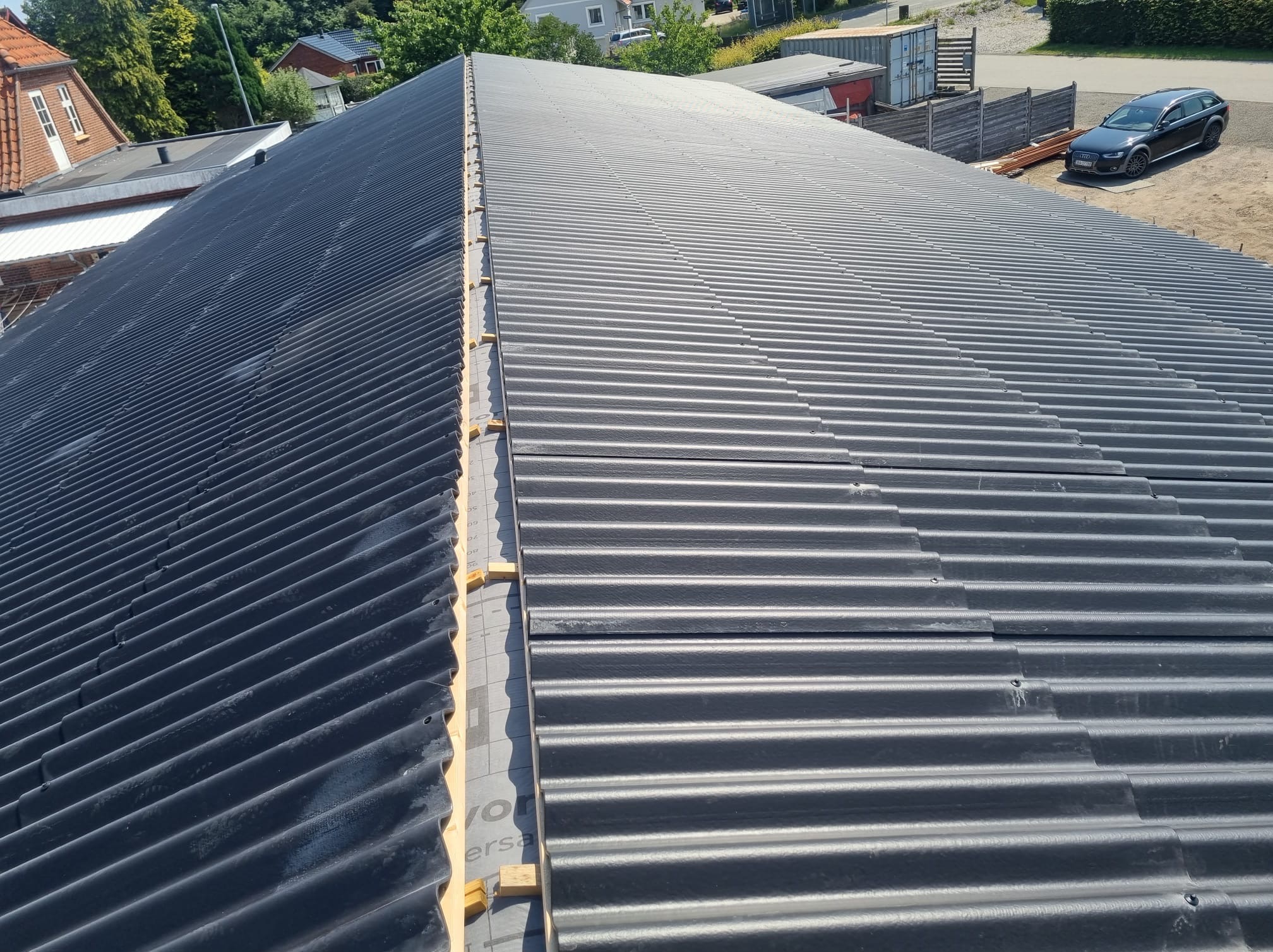 Nowe, czarne, faliste pokrycie dachowe z widoczną membraną i łatami, ujęcie z góry, w tle zabudowania i zaparkowany samochód.