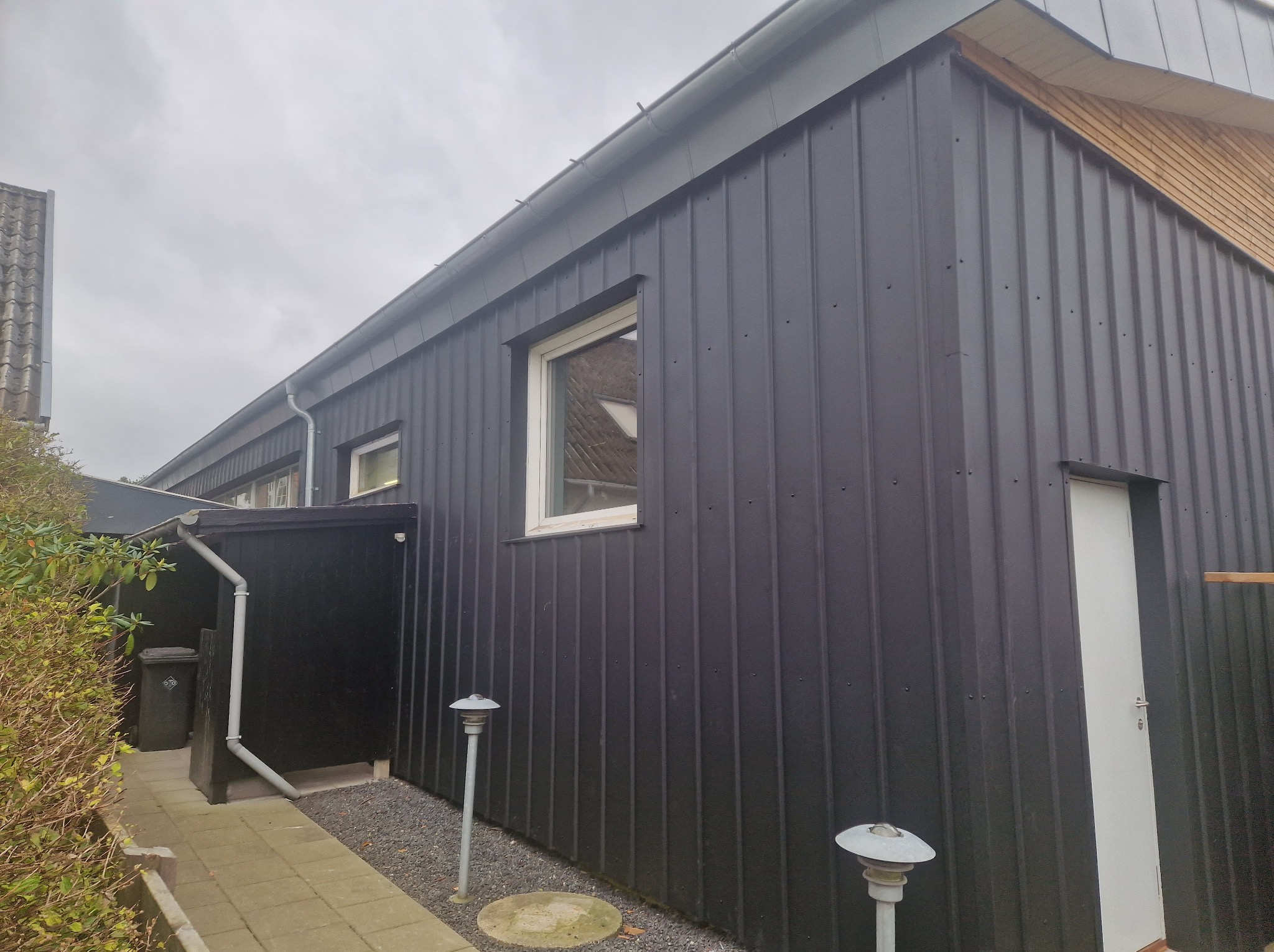 Nowoczesny budynek z ciemną, pionową blachą trapezową na elewacji, szara rynna, biała stolarka okienna i drzwiowa, oświetlenie zewnętrzne w formie lamp na słupkach.