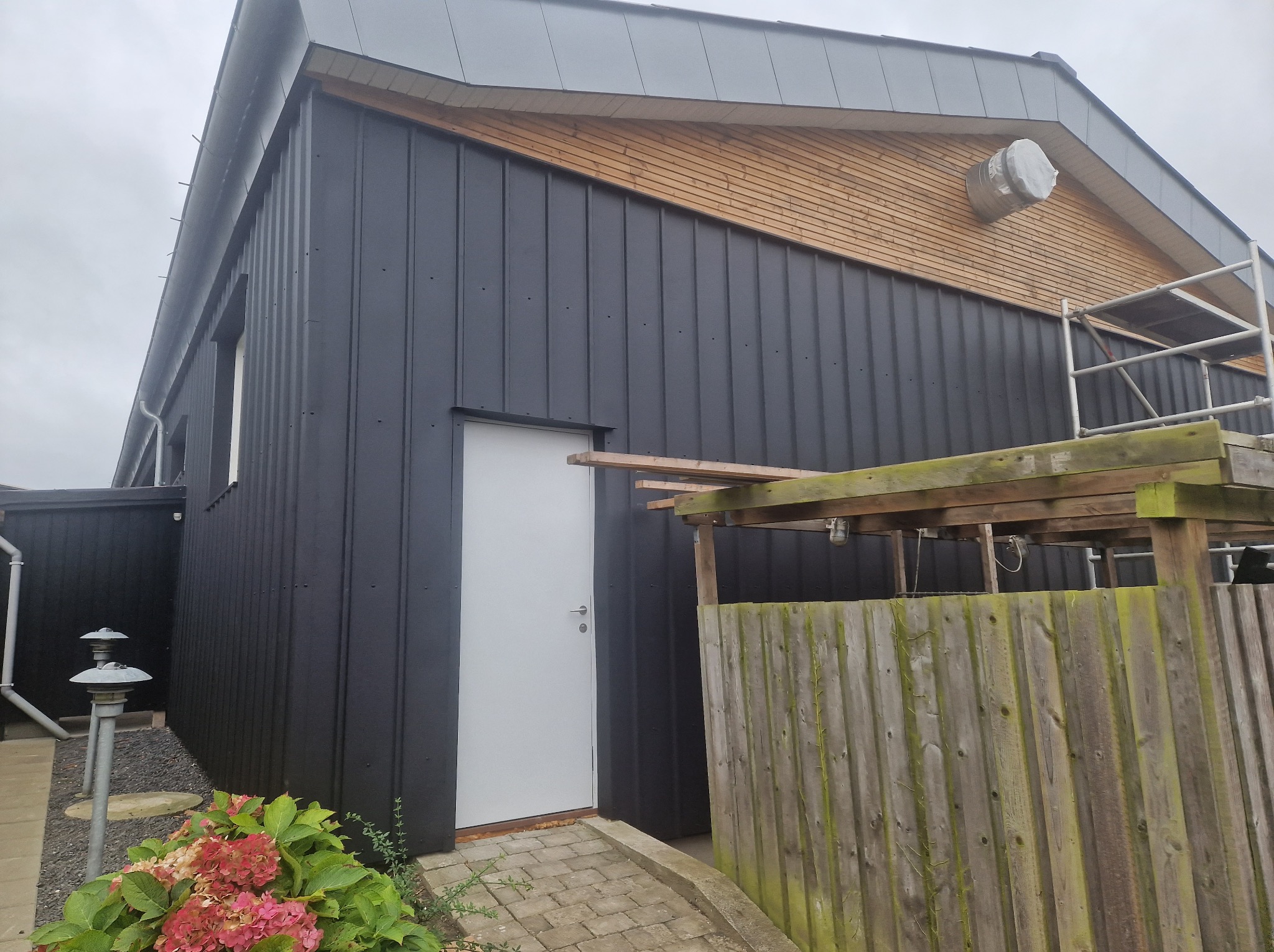 Nowoczesny dom z czarną elewacją z pionowych paneli, drewnianym akcentem pod dachem i szarą blachą dachową, widoczne rusztowanie i fragment zarośniętego płotu.