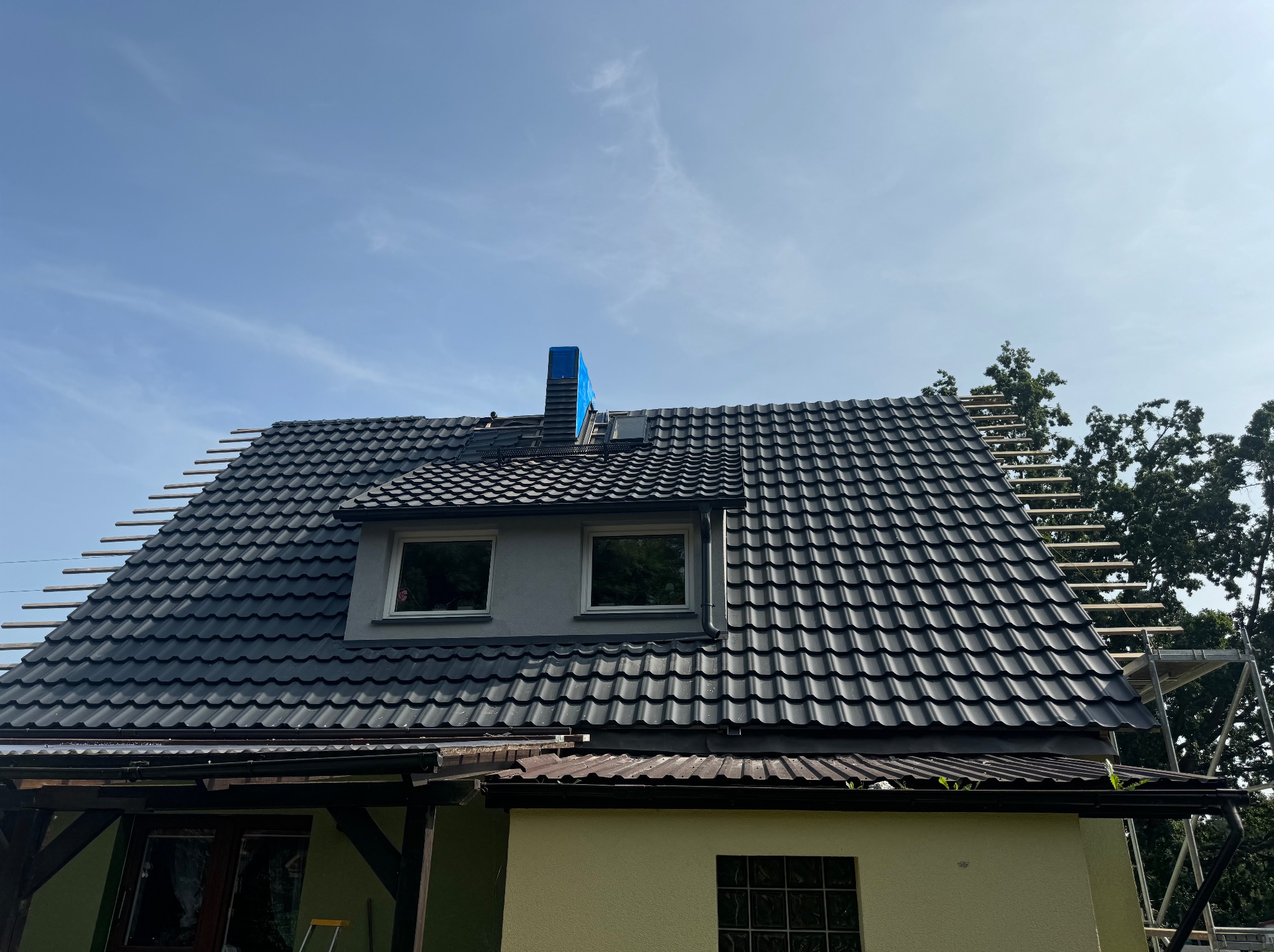 Częściowo pokryty dach z czarnej dachówki, widoczne deski na niezakończonej części, komin zabezpieczony niebieską folią, fragment elewacji domu w odcieniach żółci i szarości, oraz rusztowanie po...