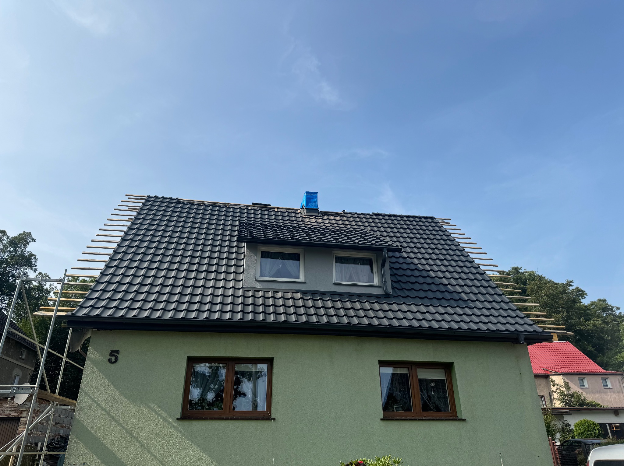 Dom z nowym, czarnym dachem z dachówki ceramicznej w trakcie montażu, widoczne łaty dachowe i zabezpieczony komin niebieską folią.