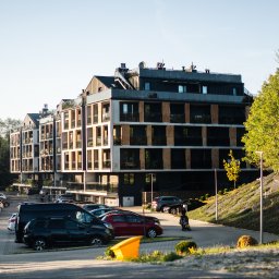 BOGDAŃSKI SPÓŁKA JAWNA JAN I CZESŁAW BOGDAŃSCY - Nowoczesny budynek apartamentowy z balkonami i dużymi oknami, otoczony zielenią i parkingiem z autami w słoneczny dzień. Widoczne detale architektoniczne.