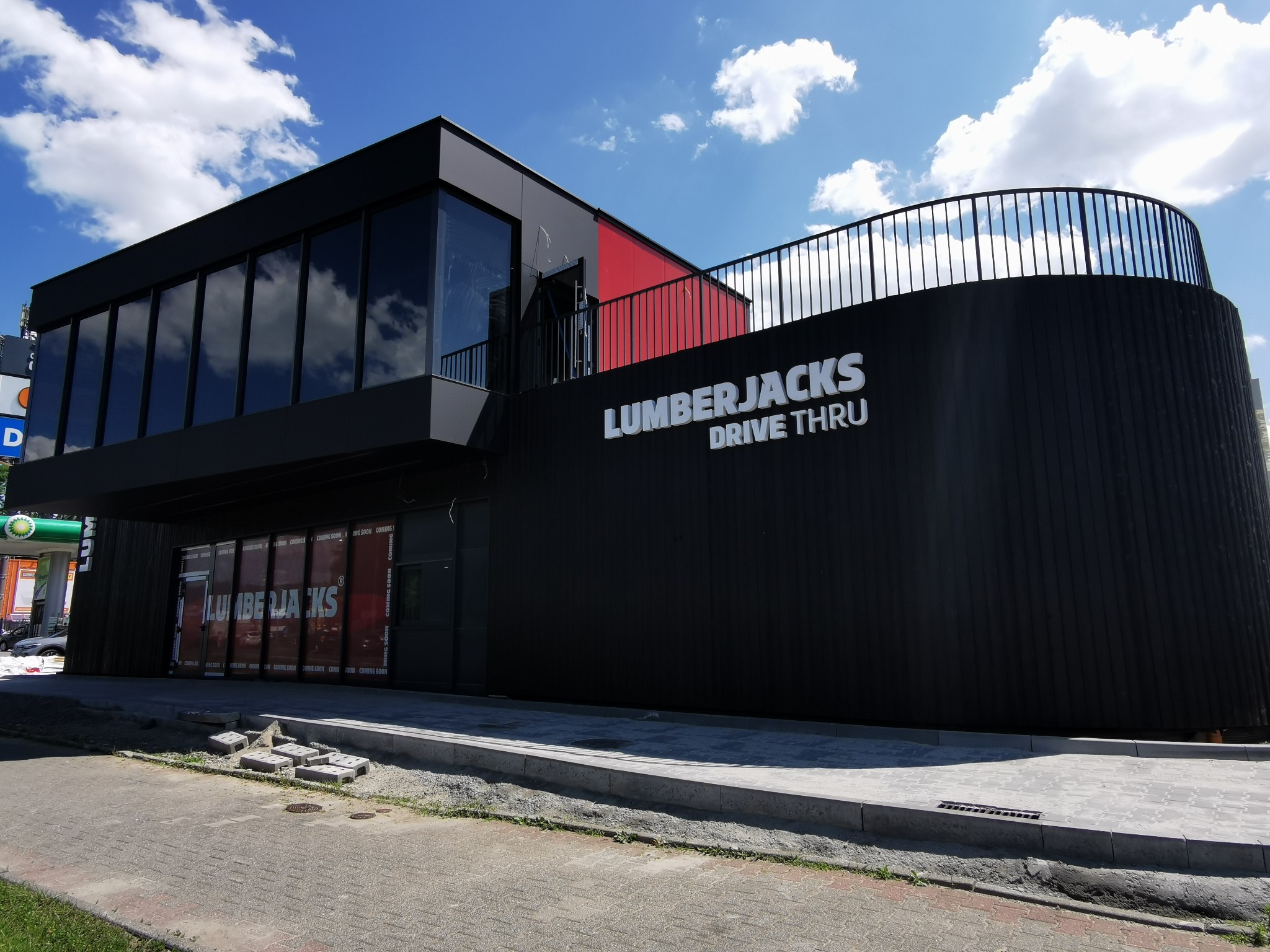 Nowoczesny budynek z dużymi oknami i zaokrągloną elewacją, na której widnieje napis 'LUMBERJACKS DRIVE THRU'. Błękitne niebo z chmurami w tle.