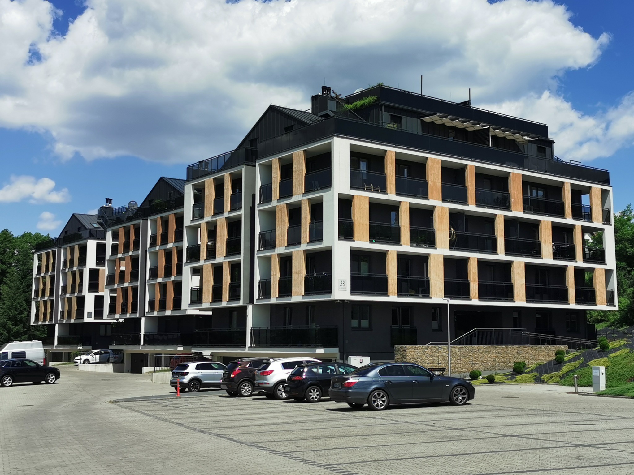 Nowoczesny budynek apartamentowy z balkonami, widok z parkingu. Architektura z elementami drewna, błękitne niebo z chmurami w tle. Elegancki design.