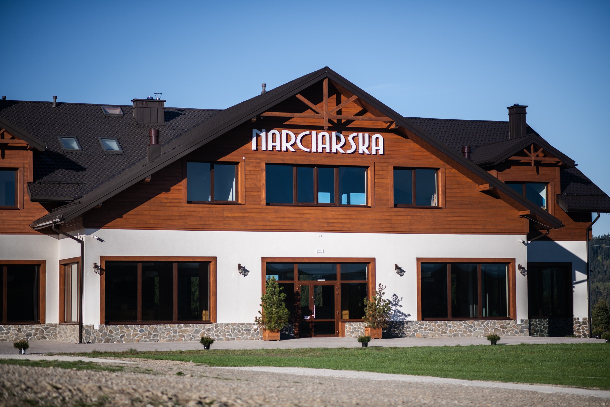 Elewacja budynku z brązowego drewna i białego tynku, z ciemnymi oknami i napisem 'NARCIARSKA' nad wejściem, podkreślona kamienną podmurówką i zielenią.