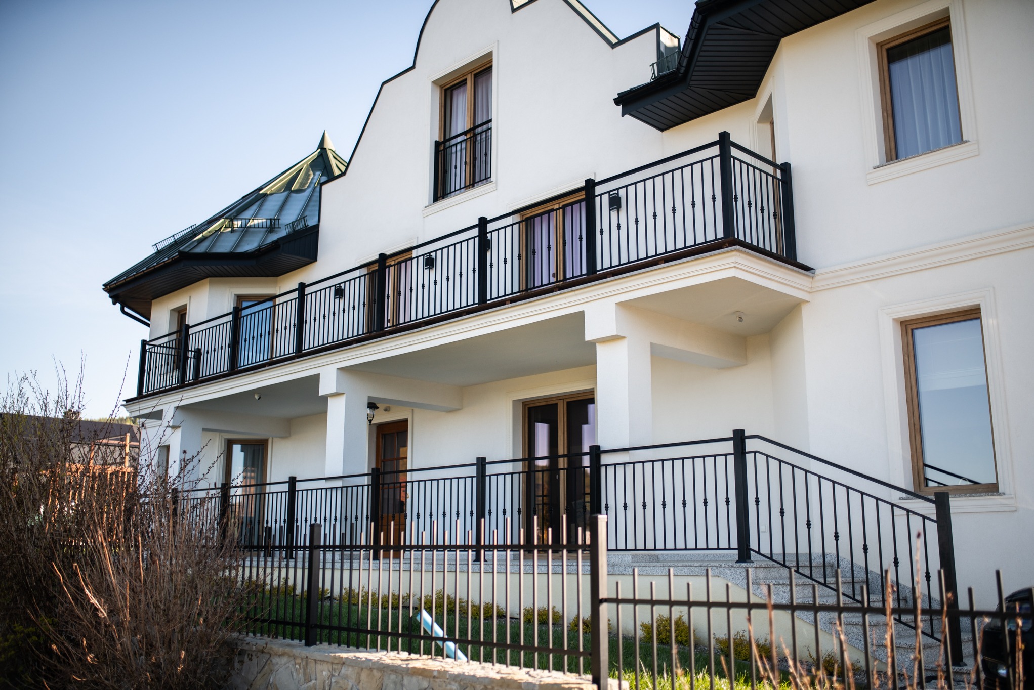 Elewacja dwupiętrowego domu z oknami w drewnianych ramach i czarnymi, kutymi balustradami balkonów w Nowym Sączu. Jasna fasada, widok z dołu, błękitne niebo.