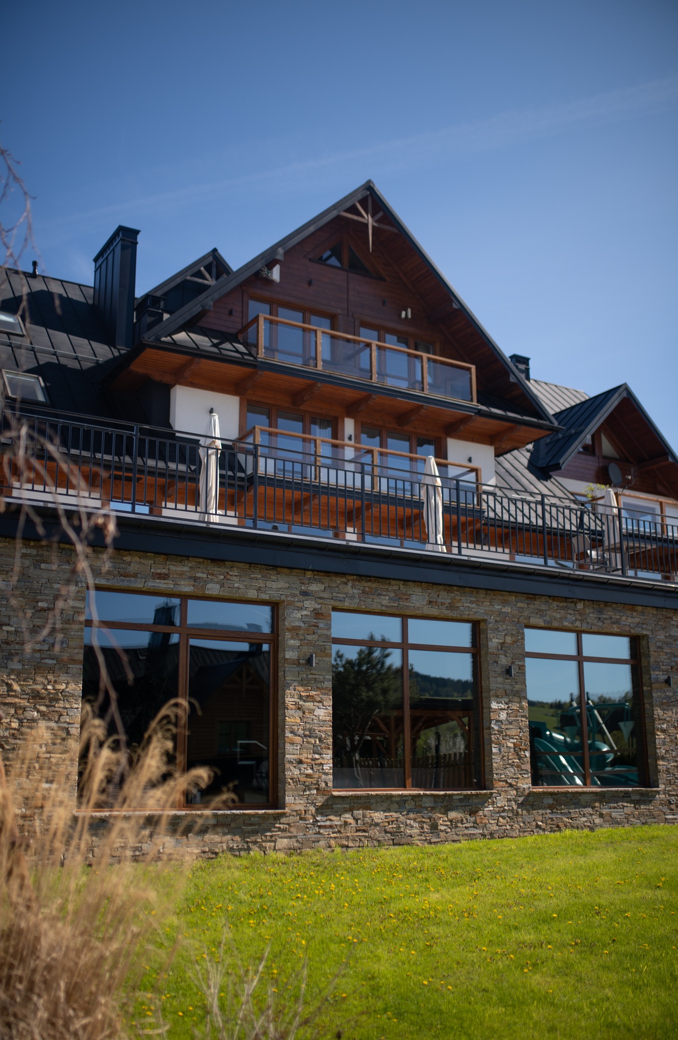 Dwukondygnacyjny dom z kamienną elewacją i drewnianymi balkonami, widok z poziomu trawnika w słoneczny dzień. W oknach odbija się otoczenie.