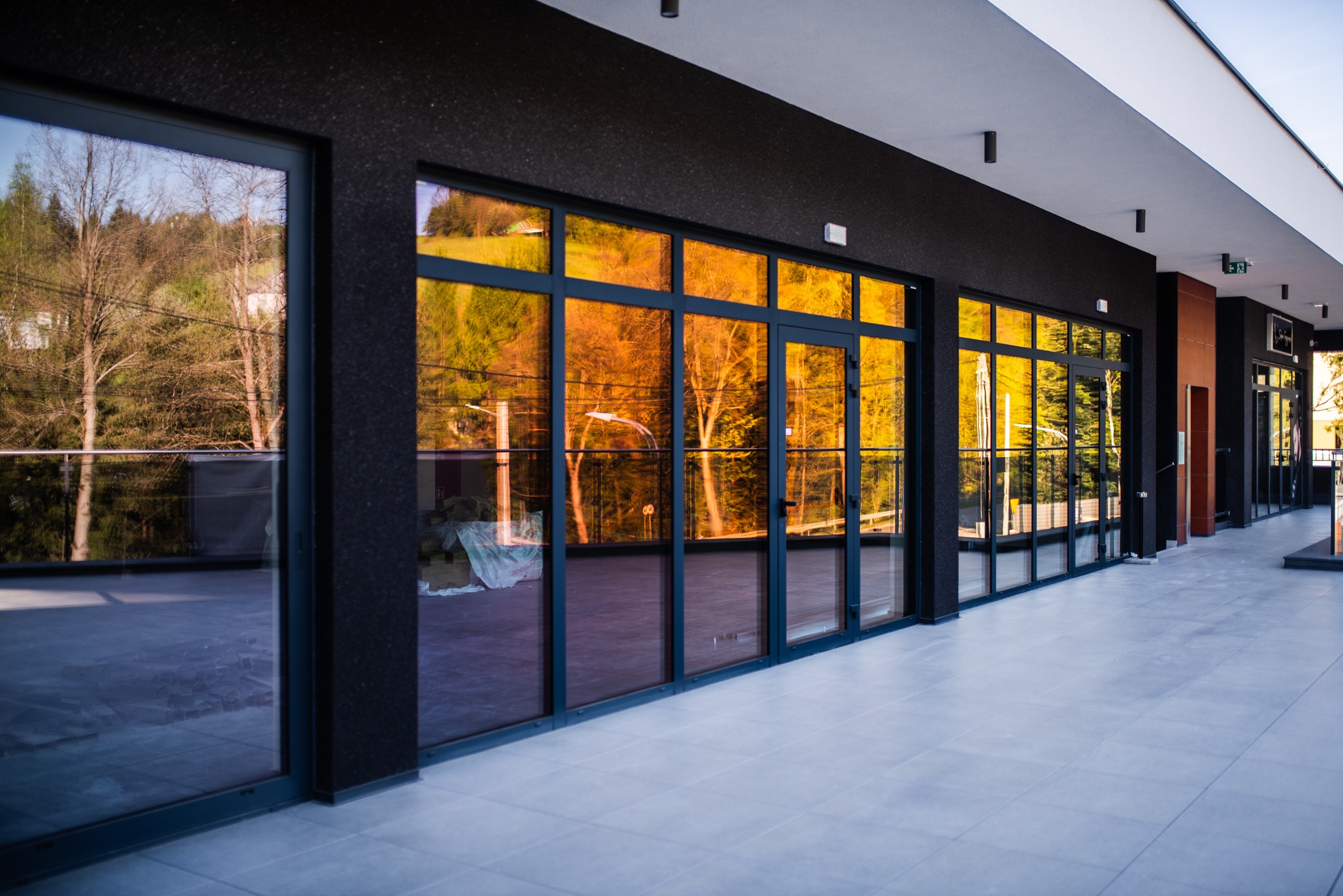 Szklana fasada budynku z ciemnymi ramami okiennymi, odbijająca krajobraz drzew. Widok z zewnątrz na taras wyłożony płytkami. Nowoczesna architektura.