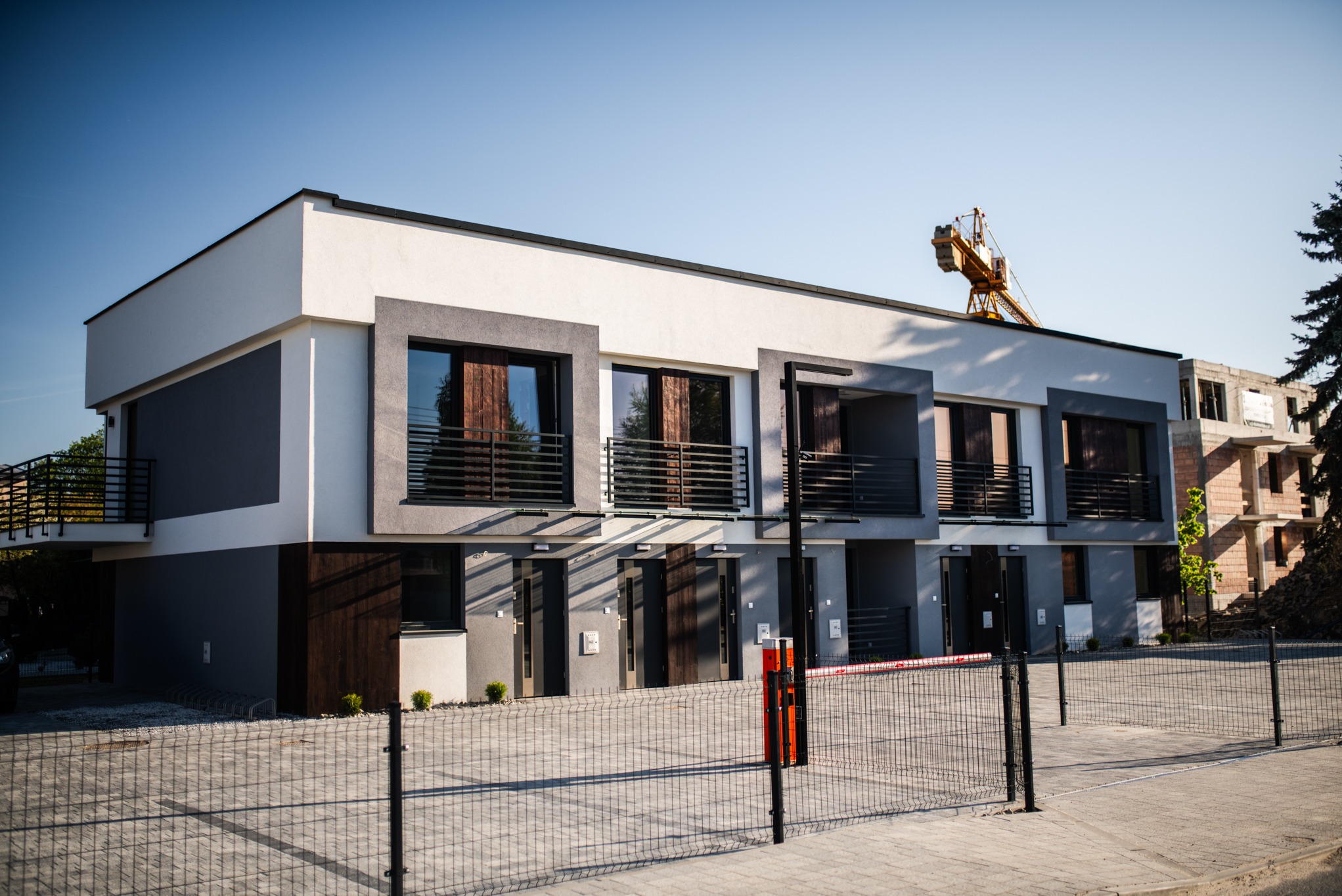 Fasada szeregowca w nowoczesnej kolorystyce z balkonami i drewnianymi elementami. W tle widoczny żuraw budowlany i budynek w trakcie budowy.