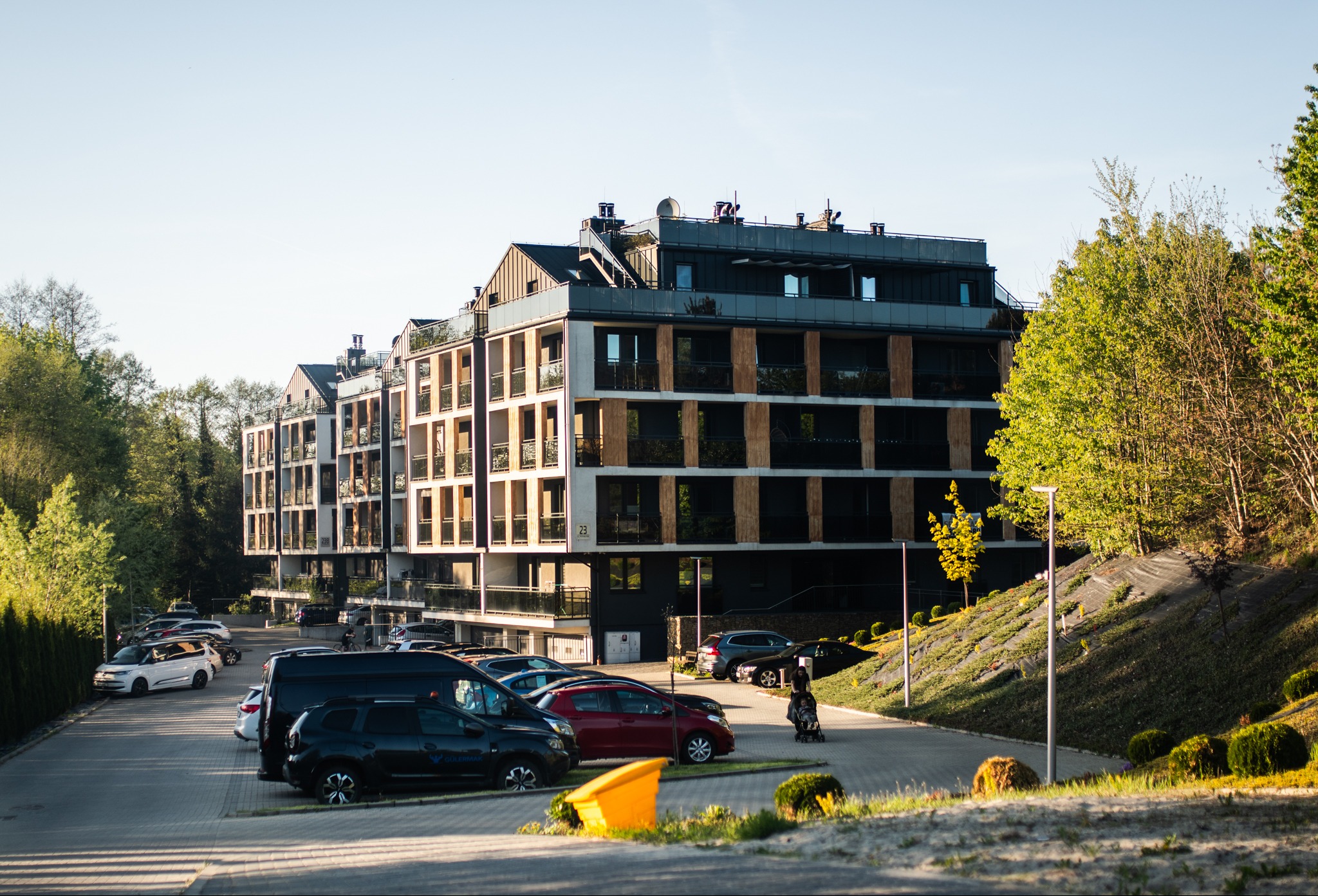 Nowoczesny budynek apartamentowy z balkonami i dużymi oknami, otoczony zielenią i parkingiem z autami w słoneczny dzień. Widoczne detale architektoniczne.