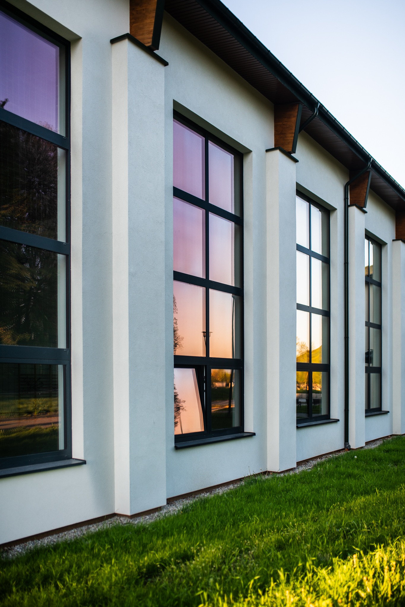 Elewacja budynku z rzędem okien z czarnymi ramami, w których odbija się zachód słońca, podkreślona przez pionowe elementy architektoniczne.