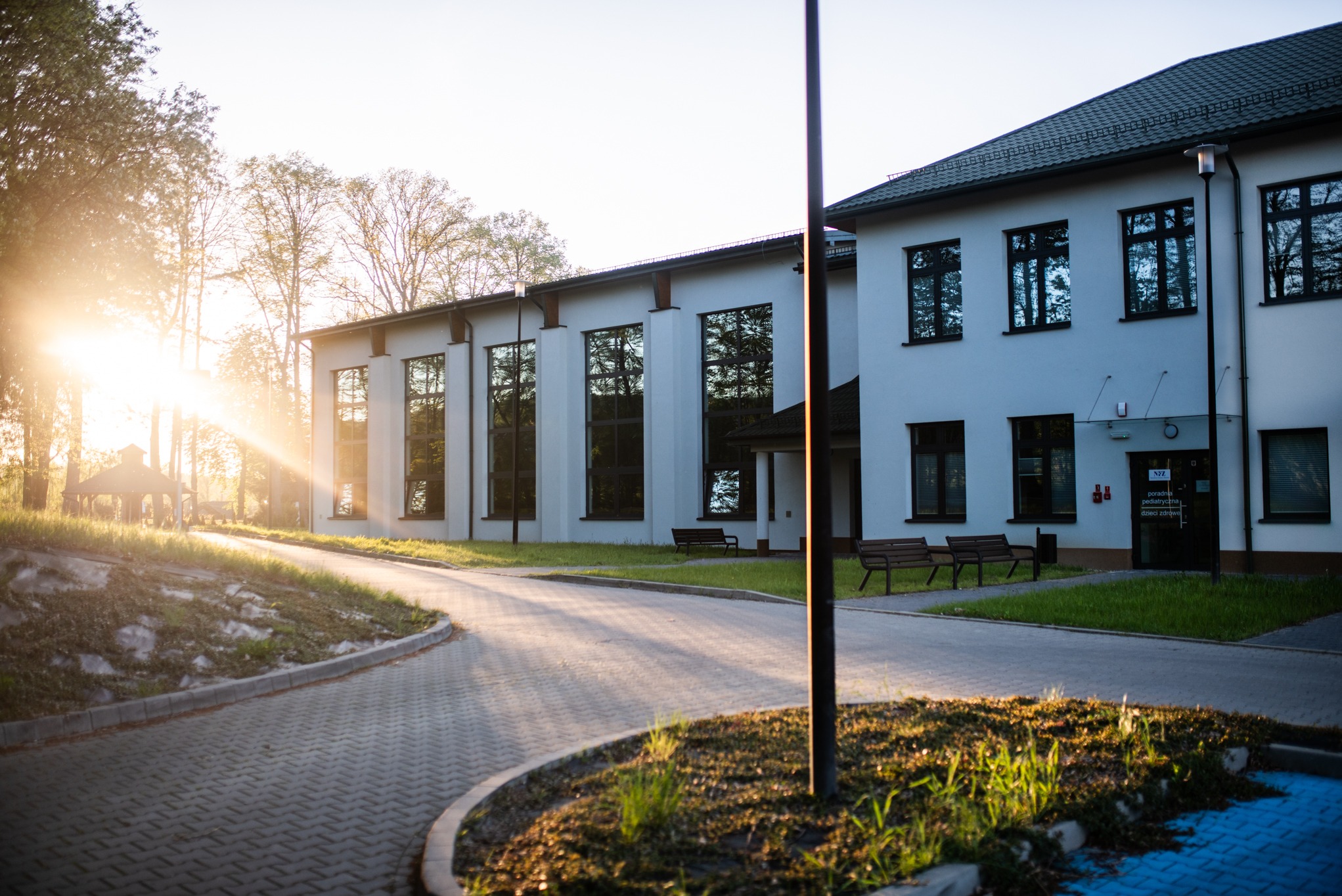 Elewacja budynku z dużymi oknami, odbijającymi drzewa. Promienie słoneczne oświetlają fasadę i chodnik, tworząc ciepłą atmosferę. Widoczne ławki i zadbana zieleń.