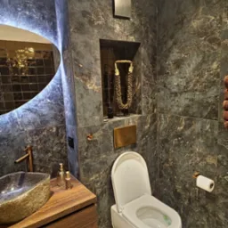 Elegancka łazienka z ciemnymi marmurkowymi płytkami, podświetlanym lustrem o nieregularnym kształcie, umywalką z kamienia na drewnianej szafce, wiszącą toaletą i złotymi akcentami.
