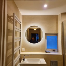 Klusbedrijf Ceasar - Nowoczesna łazienka w beżowych płytkach z okrągłym lustrem podświetlanym LED, białą umywalką nablatową na czarnej szafce, grzejnikiem drabinkowym i oknem z akcesoriami.