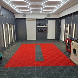 Klusbedrijf Ceasar - Wnętrze garażu lub warsztatu z szarą ścianą, jasnym oświetleniem sufitowym w kształcie plastra miodu i podłogą z czerwonych i szarych paneli. Widoczna pralka i wózek z narzędziami.