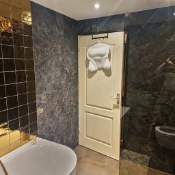 Klusbedrijf Ceasar - Luksusowa łazienka z czarno-złotymi płytkami, marmurowymi ścianami, białą wanną i poduszką na drzwiach.