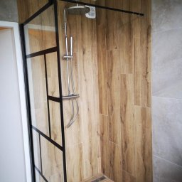 Klusbedrijf Ceasar - Kabina prysznicowa walk-in z czarnymi profilami, wykończona płytkami imitującymi drewno i szarym gresem, z odpływem liniowym w podłodze.