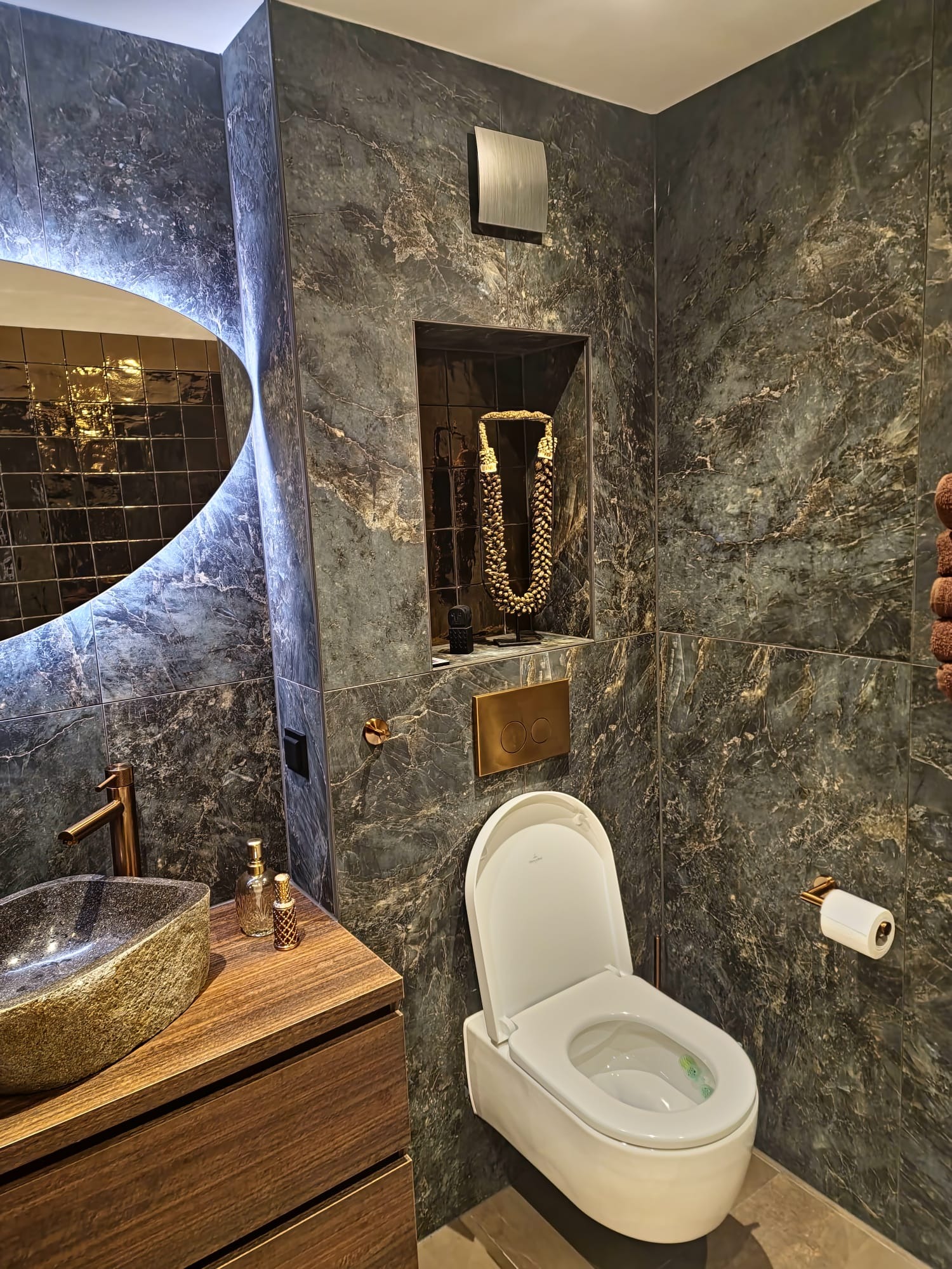Elegancka łazienka z ciemnymi marmurkowymi płytkami, podświetlanym lustrem o nieregularnym kształcie, umywalką z kamienia na drewnianej szafce, wiszącą toaletą i złotymi akcentami.