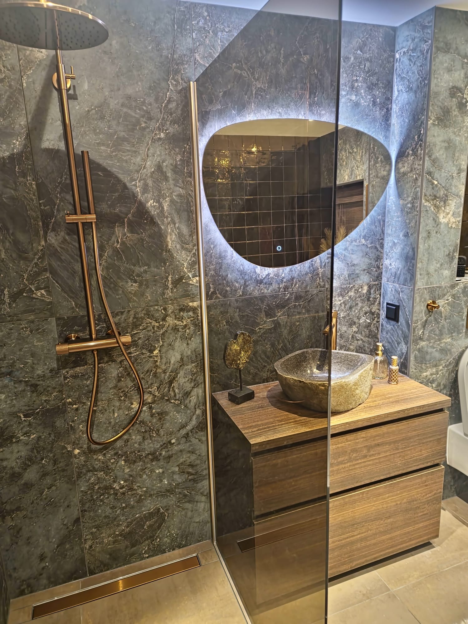 Nowoczesna aranżacja łazienki w Świętajnie z marmurowymi płytkami, deszczownicą w kolorze miedzi, umywalką z kamienia i podświetlanym lustrem o nieregularnym kształcie.