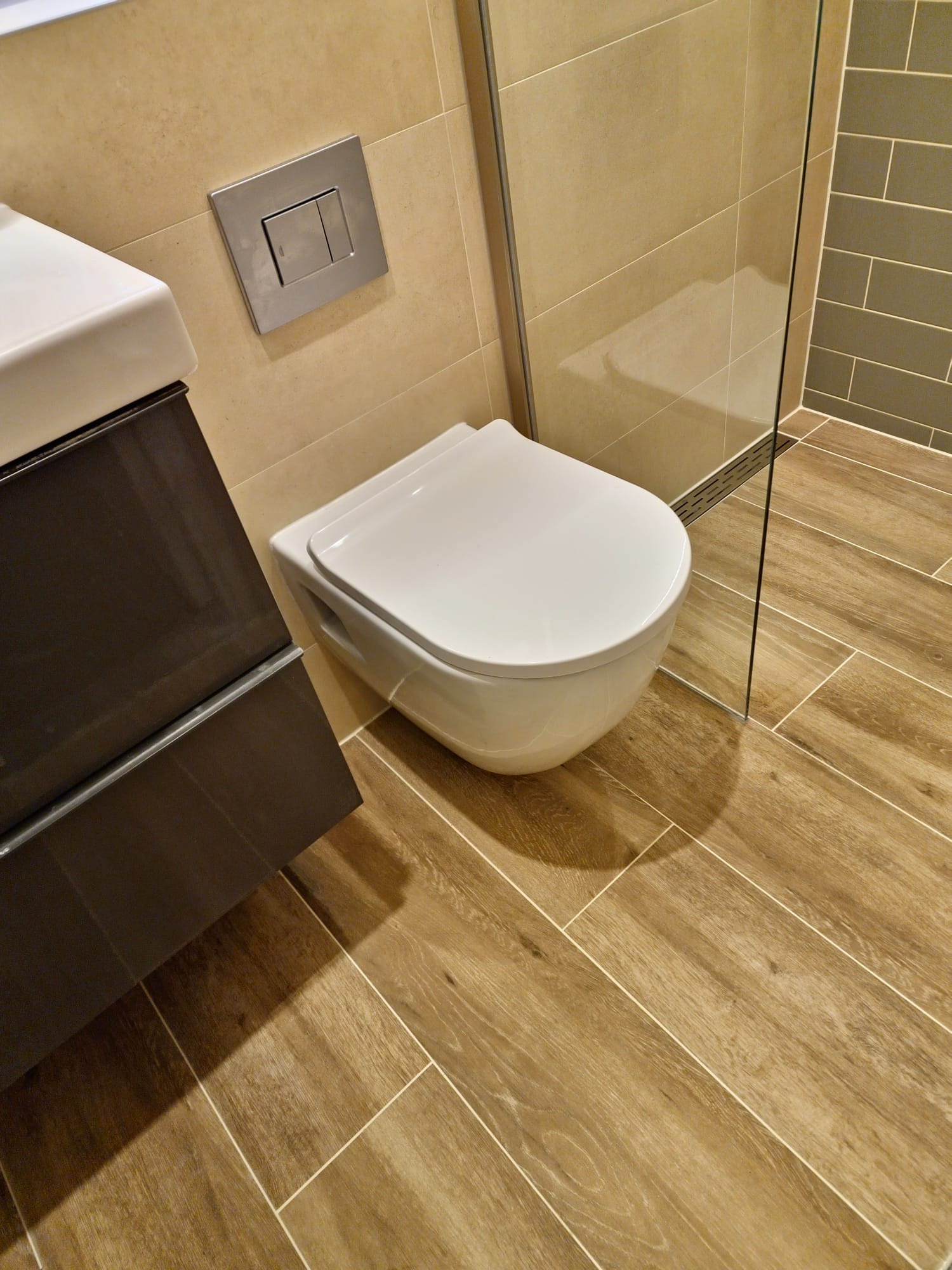 Nowoczesna łazienka z podwieszaną toaletą, drewnopodobnymi płytkami na podłodze i ścianie, oraz szklaną kabiną prysznicową z odpływem liniowym.