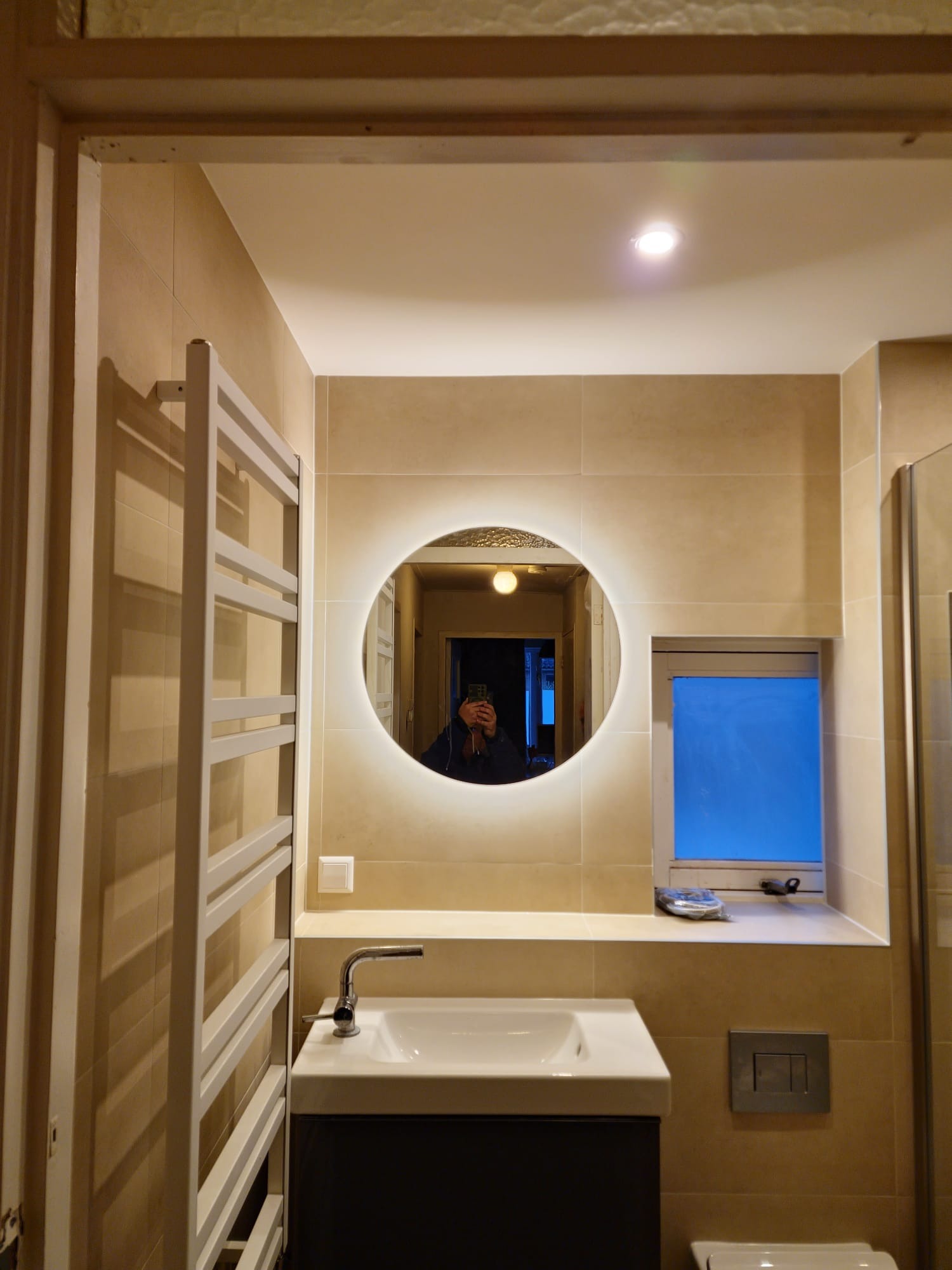 Nowoczesna łazienka w beżowych płytkach z okrągłym lustrem podświetlanym LED, białą umywalką nablatową na czarnej szafce, grzejnikiem drabinkowym i oknem z akcesoriami.