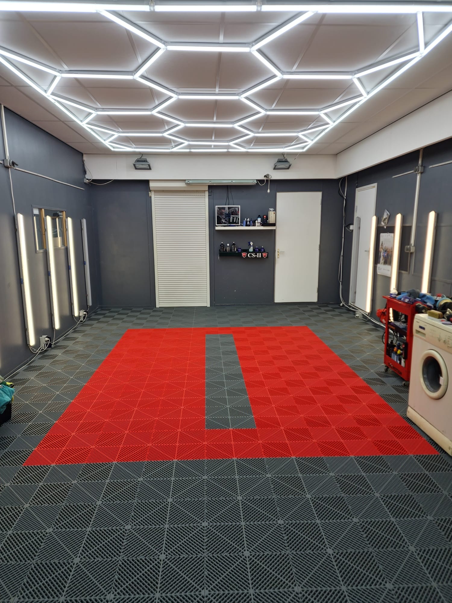 Wnętrze garażu lub warsztatu z szarą ścianą, jasnym oświetleniem sufitowym w kształcie plastra miodu i podłogą z czerwonych i szarych paneli. Widoczna pralka i wózek z narzędziami.