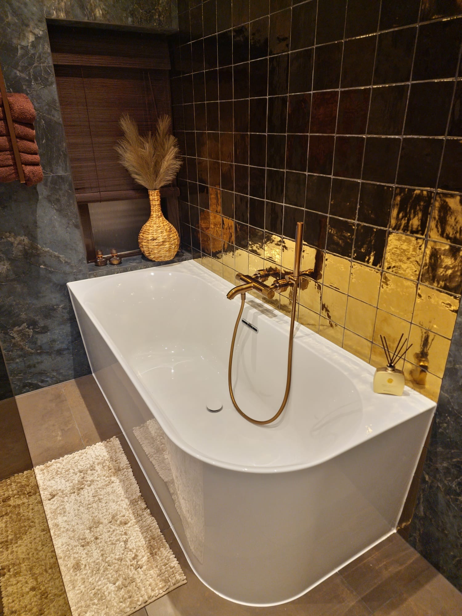 Nowoczesna łazienka z białą wanną narożną, złotą armaturą i dekoracyjną ścianą z płytek w odcieniach czerni i złota, uzupełniona o pleciony wazon z suszonymi trawami i dyfuzor zapachowy.