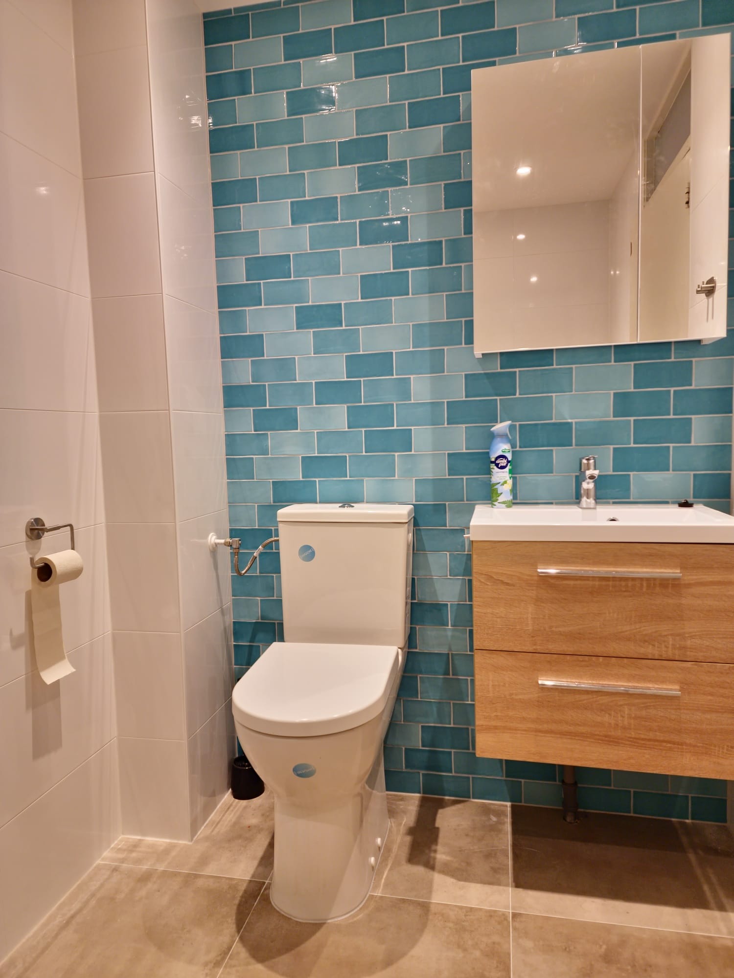 Wykończona łazienka z toaletą, umywalką z drewnianą szafką i lustrem, ściany wyłożone turkusowymi płytkami cegiełkami i białymi płytkami.