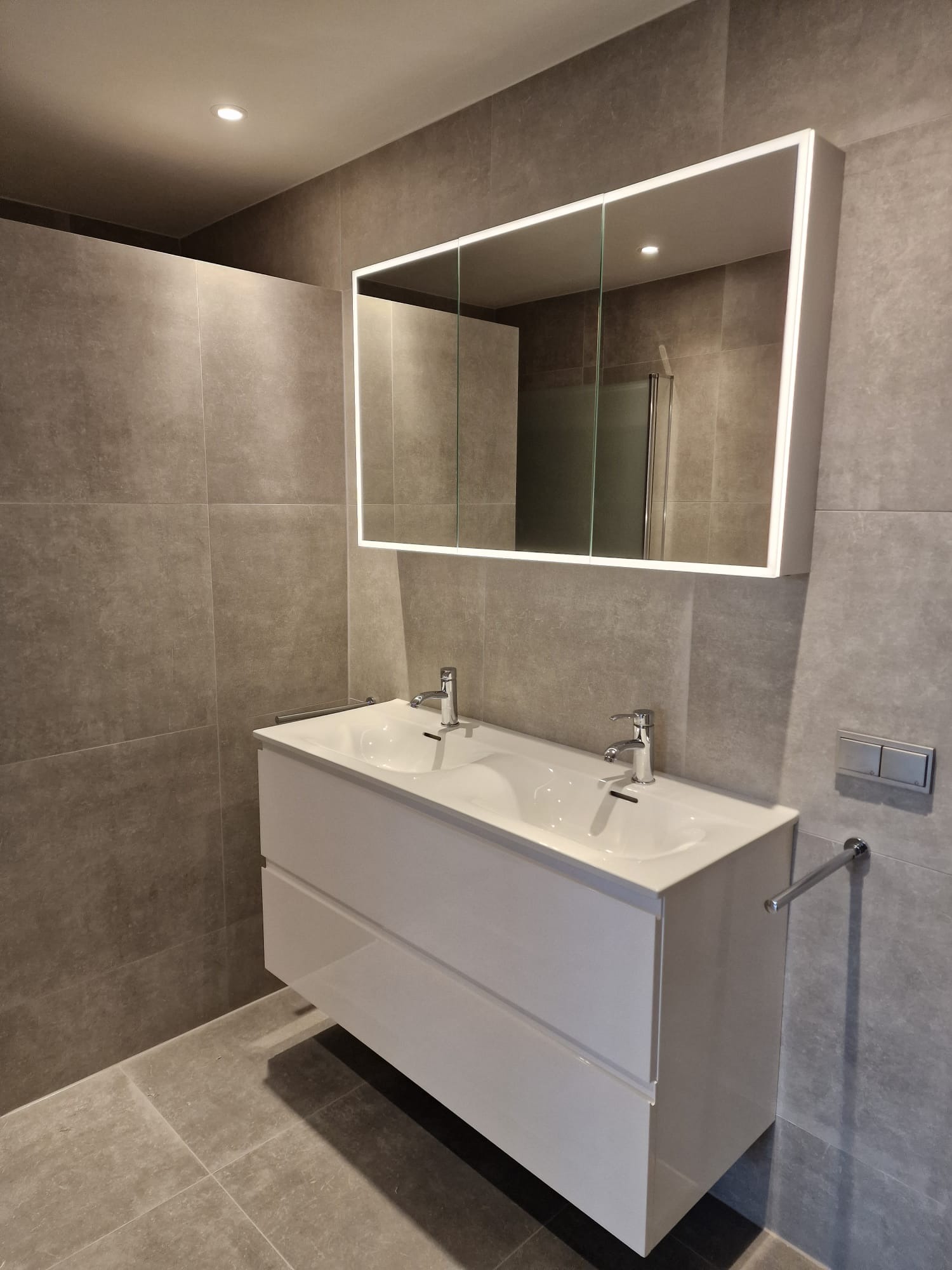 Nowoczesna łazienka z szarymi płytkami, białą szafką pod umywalką z dwoma umywalkami i podświetlanym lustrem z szafką.