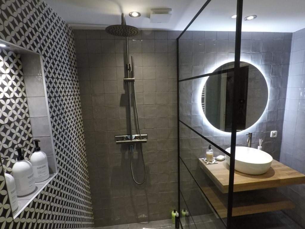 Nowoczesna łazienka w odcieniach szarości z kabiną prysznicową typu walk-in, okrągłym lustrem z podświetleniem LED i umywalką nablatową na drewnianym blacie.