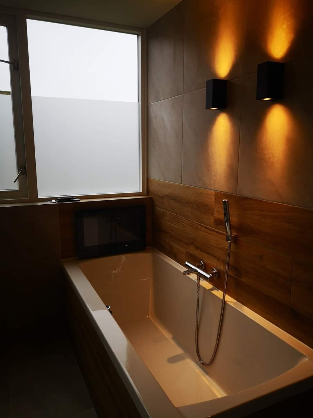 Nowoczesna łazienka z wanną, telewizorem w zabudowie, oświetleniem ściennym i oknem z częściowo matową szybą.