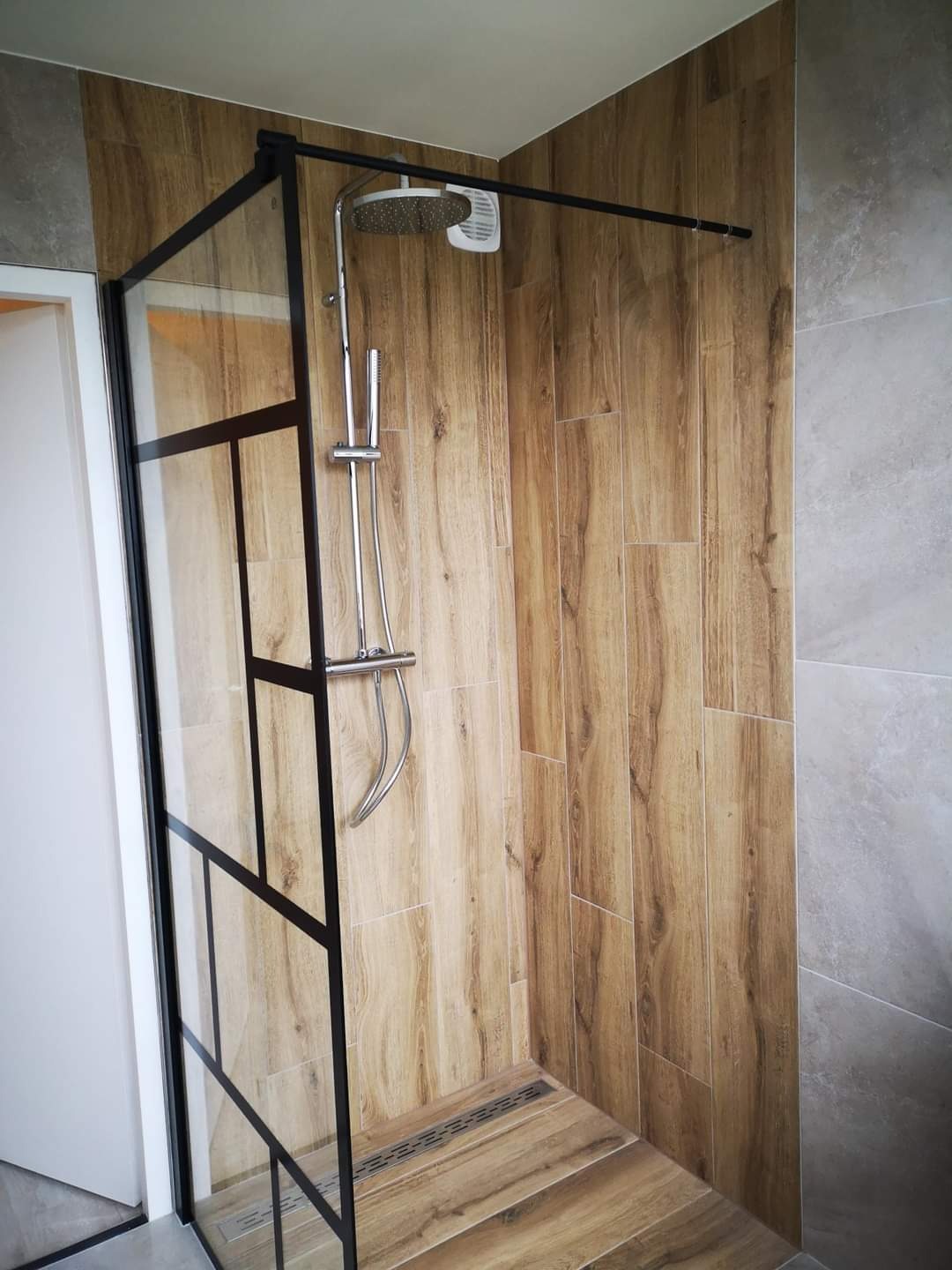Kabina prysznicowa walk-in z czarnymi profilami, wykończona płytkami imitującymi drewno i szarym gresem, z odpływem liniowym w podłodze.
