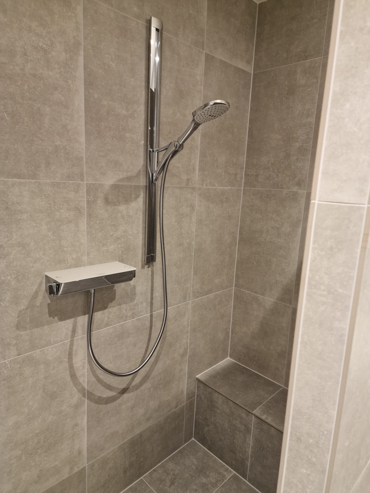 Wykończona kabina prysznicowa z szarymi, matowymi płytkami, chromowaną armaturą i wbudowanym siedziskiem.