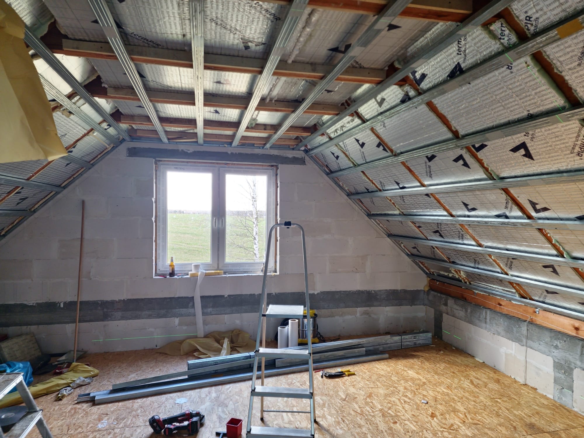 Poddasze w trakcie remontu: ocieplony dach z widoczną konstrukcją metalową, ściany z bloczków, okno z widokiem na zielone pole, drabina i narzędzia na podłodze z płyt OSB.
